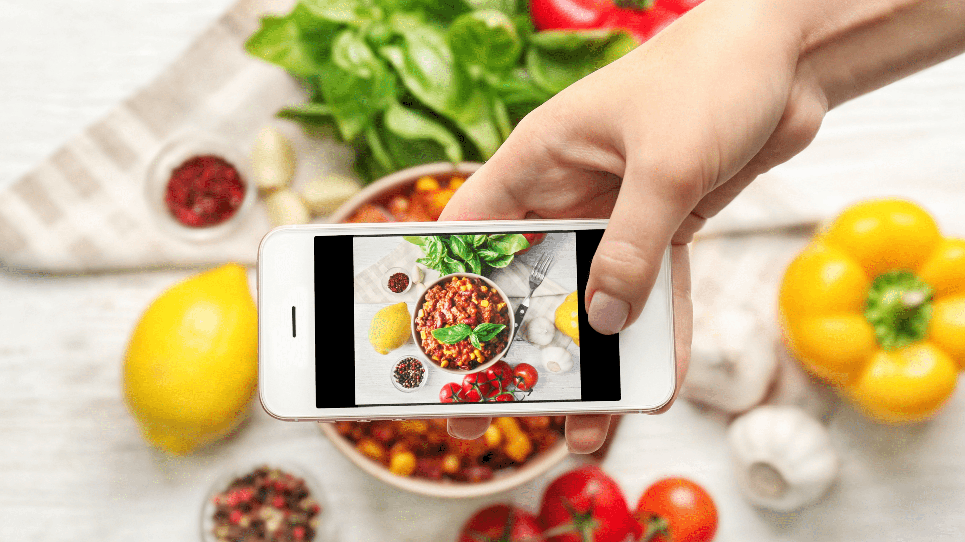 person's hand with smartphone taking a picture of a bowl of pasta with a yellow bell pepper, lemon, tomatoes, garlic, basil, black peppers, and fork surrounding it