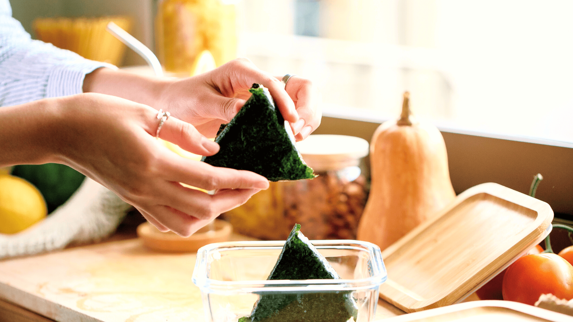 personne plaçant des onigiri triangulaires dans un Tupperware en verre avec un autre onigiri triangulaire et une courge musquée en arrière-plan