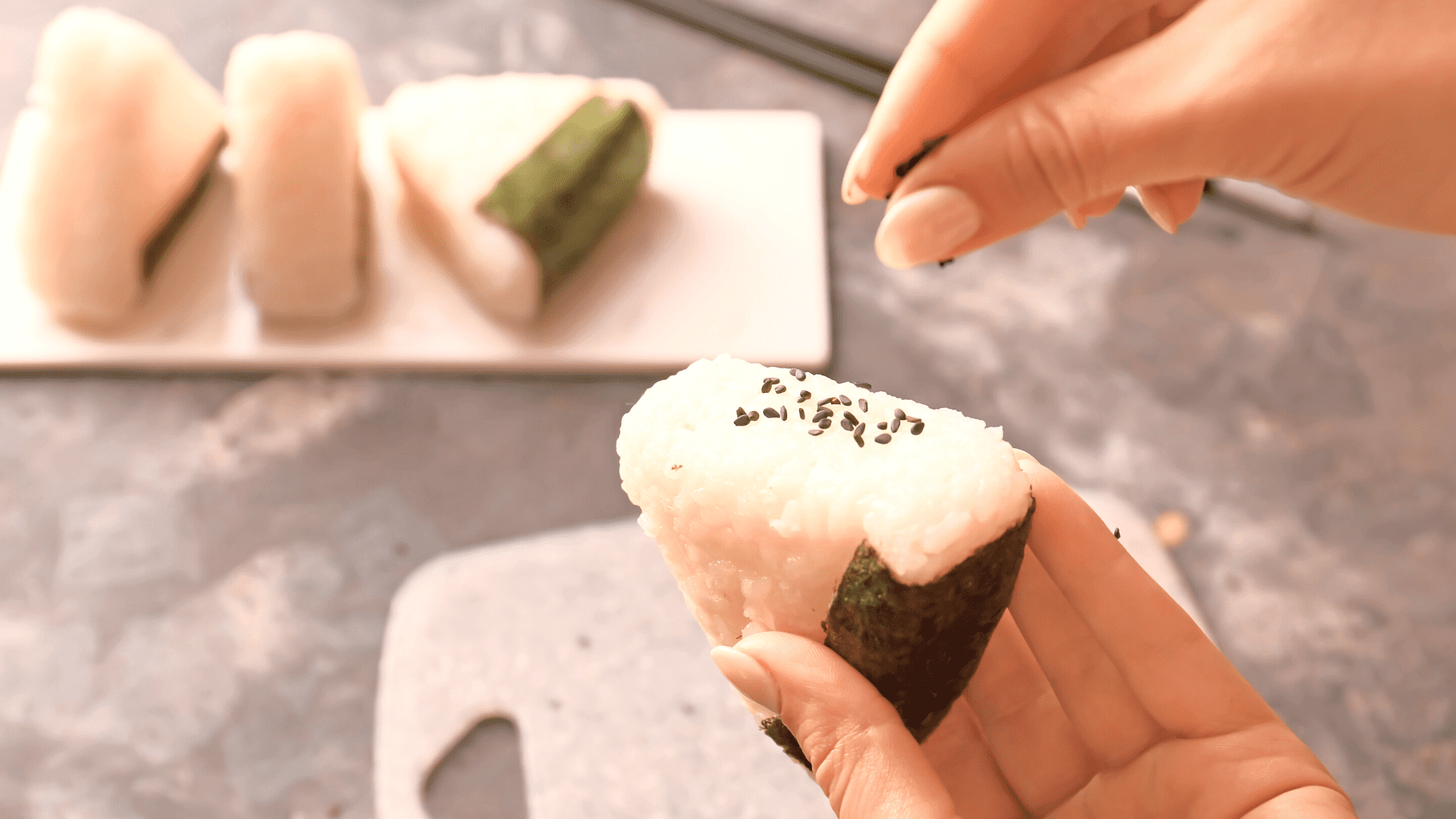 Primer plano de alguien rociando sésamo negro sobre onigiri triangular con 3 en el fondo