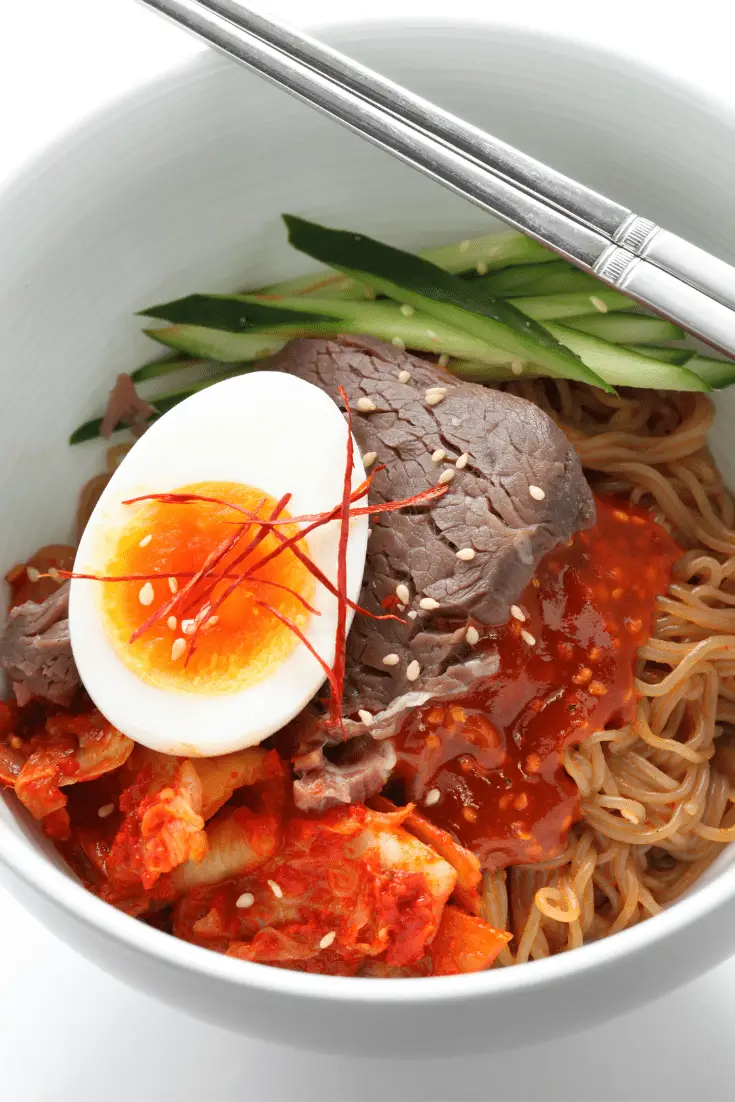 white boil of naengmyeon with boiled egg, beef, and cucumber, with silver chopsticks