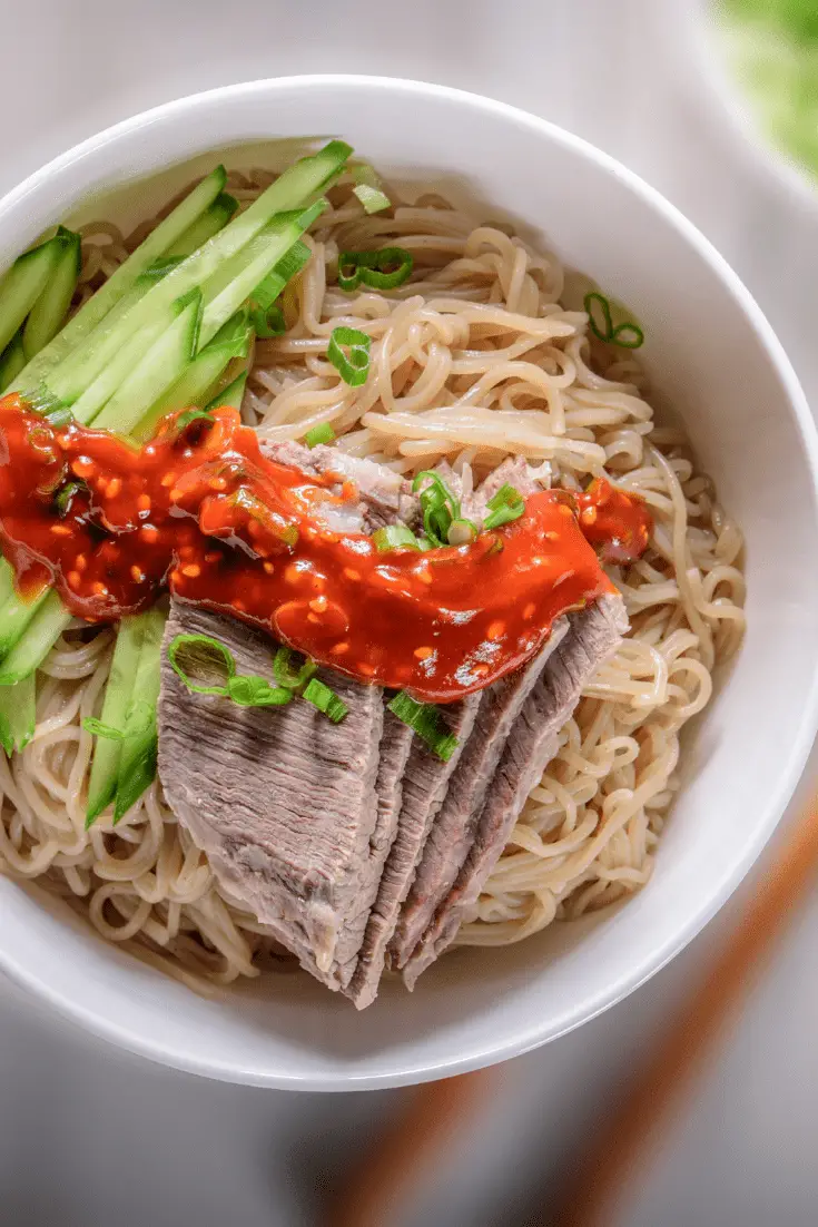 white bowl of naengmyeon with beef, cucumber, green onion, and hot sauce