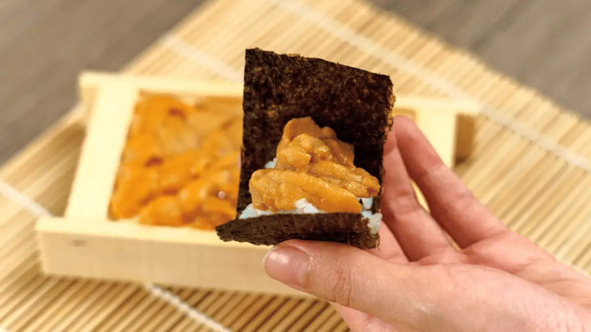 hand holding seaweed with rice and uni inside, container of uni in the background